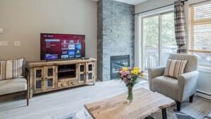 Una sala de estar con una televisión de pantalla plana, una chimenea de piedra, un mueble de entretenimiento de madera, un sillón gris y una mesa de centro con un jarrón de flores.