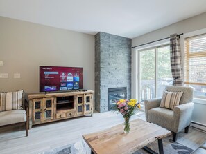 Ein Wohnzimmer mit Flachbildfernseher, Kamin aus Stein, Holzunterhaltungsmöbel, ein grauer Sessel und ein Couchtisch mit Blumenvase.