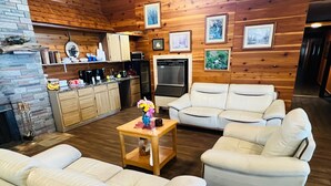 A cozy living room with a fireplace, white leather sofas, a wooden coffee table, and a kitchen area with wooden cabinets and a refrigerator.