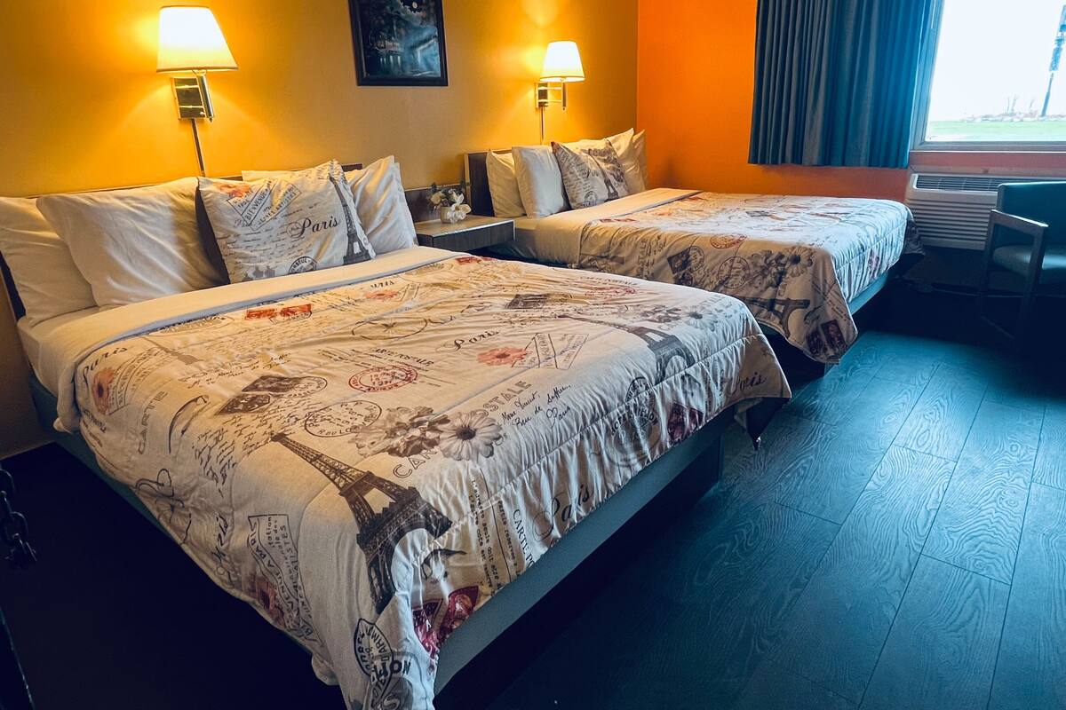A hotel room with two beds, each with a Paris-themed bedspread, two wall-mounted lamps, a painting, and a window with curtains.