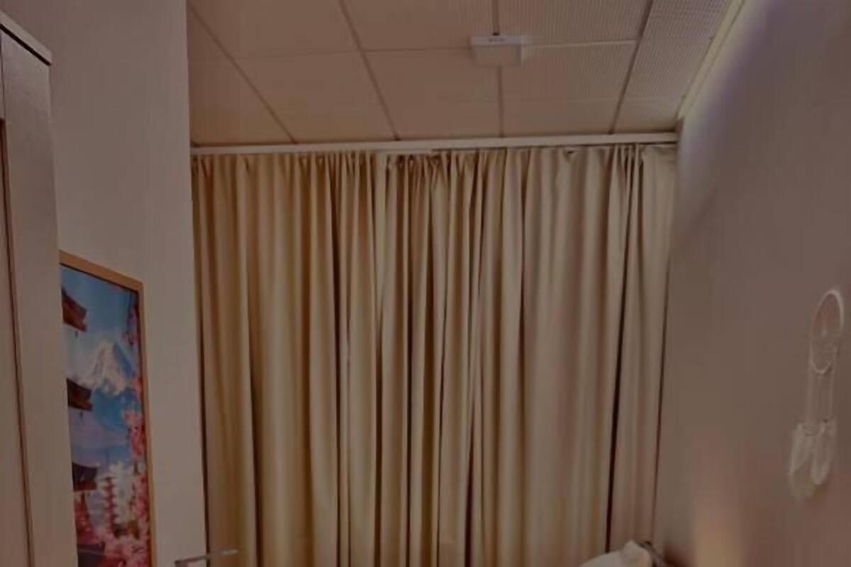 A compact hotel room with a bed, a desk, a chair, a small table, a lamp, and a drying rack.