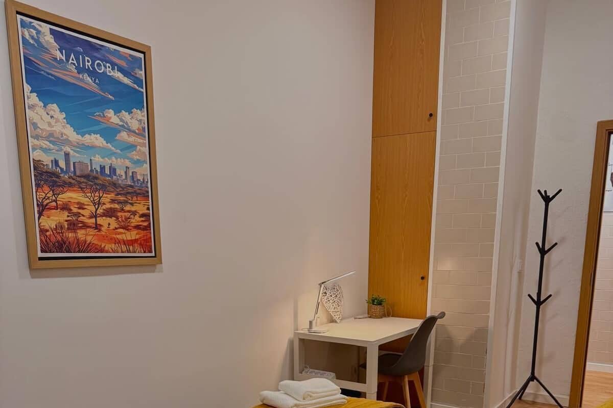 A room with two beds, a desk, a chair, a coat rack, and a framed picture of Nairobi.
