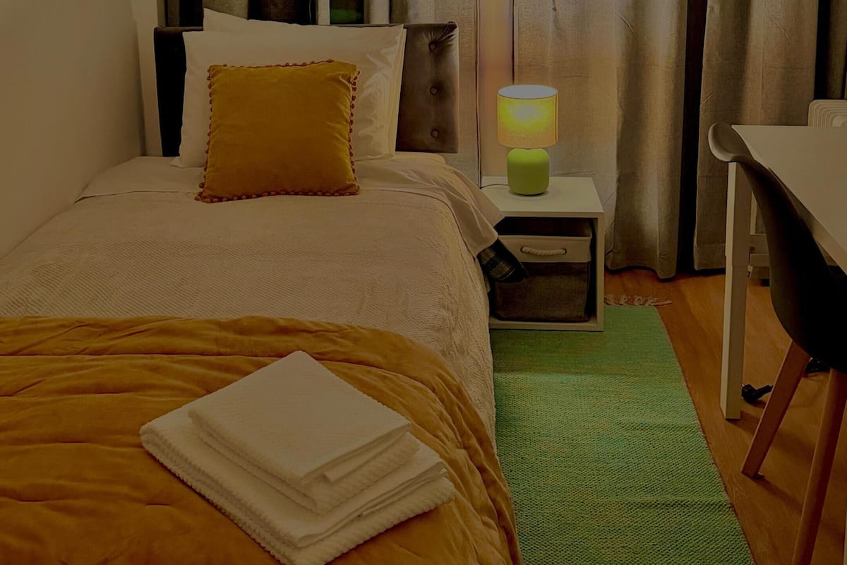 A hotel room with a bed, a nightstand, a lamp, a chair, and a view through the curtains.