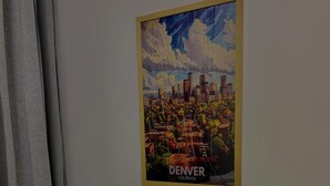 Una habitación con un escritorio blanco, una silla negra, un radiador, una cortina gris y un cuadro enmarcado de Denver.