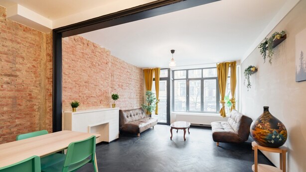 Una sala de estar moderna con pared de ladrillo, un sofá, una mesa de comedor con sillas y un jarrón grande sobre una mesita auxiliar.