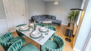 A dining area with a white table set for four, green upholstered chairs, a grey sofa in the background, and a floor lamp. - Stylish Home | Families | Groups | Contractors (Cheshire West and Chester)