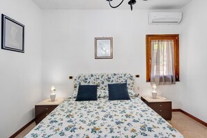 Quarto com edredom estampado de flores, duas mesas de cabeceira com luminárias, um quadro na parede e um ar condicionado.