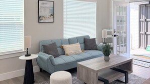Ein modernes Wohnzimmer mit einer Couch, einem Couchtisch und einem kleinen runden Hocker. Ein Fenster mit Jalousien, eine Wand-Klimaanlage und eine Tür, die nach draußen führt, sind ebenfalls zu sehen.