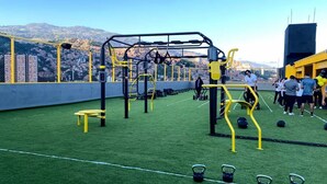 Ein Outdoor-Fitnessbereich mit Trainingsgeräten, einer grünen Rasenfläche und einem Blick auf die Stadt im Hintergrund.