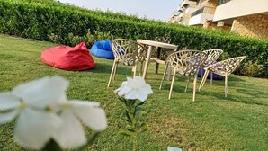 Ein grüner Außenbereich mit einem Tisch, Stühlen, einem roten Sitzsack und einer Hecke im Hintergrund.