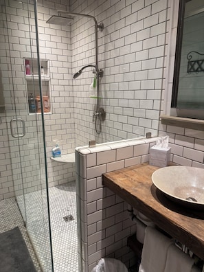 A bathroom with white tiled walls, a glass shower enclosure, a wooden vanity, and a round sink.