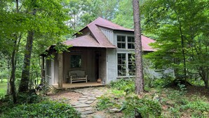 A rustic cabin surrounded by dense forest, with a stone pathway leading to the entrance.
