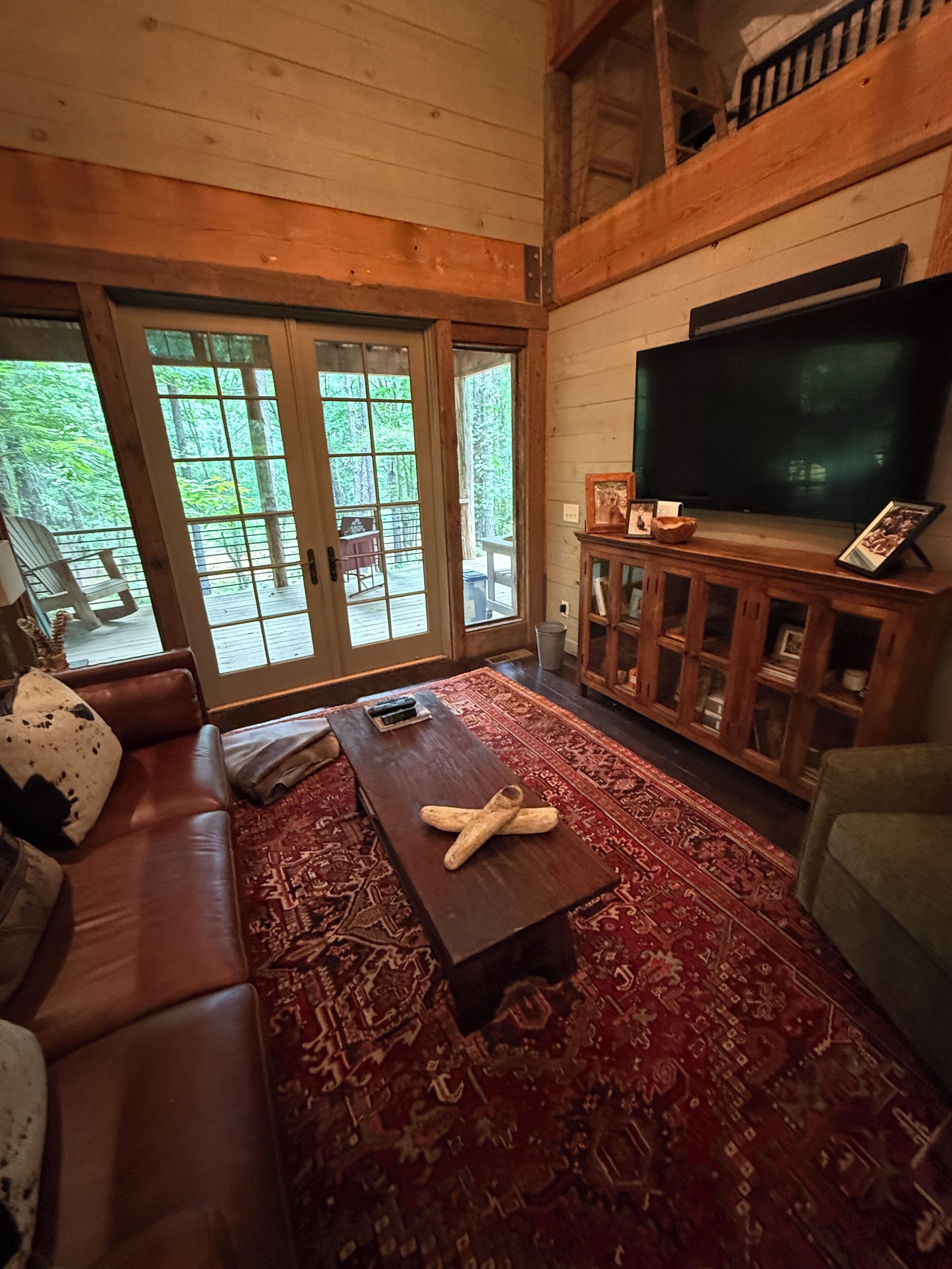 A cozy living room with a leather sofa, a coffee table, a TV, and a large window with a view of trees.