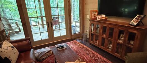 A cozy living room with a leather sofa, a coffee table, a TV, and a large window with a view of trees.