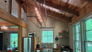 A rustic kitchen with wooden beams, a stainless steel refrigerator, a stove, and a window overlooking greenery.