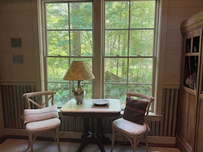 Een kamer met een groot raam, twee stoelen, een tafel met een lamp en een houten kast.