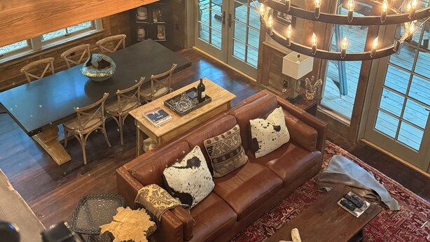 A spacious loft-style living area with a large wooden ceiling, a circular chandelier, a brown leather sofa, and a dining table with chairs.