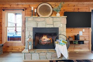 Ein Steinkamin mit Feuer, ein Fernseher an der Wand und ein Fenster mit Blick auf eine Terrasse.