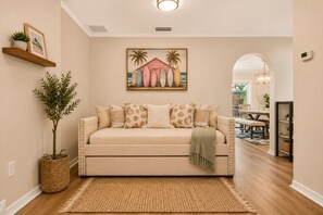 Un salon avec un canapé beige, des coussins décoratifs, un tapis tissé, un tableau accroché au mur et, au fond, un coin repas.
