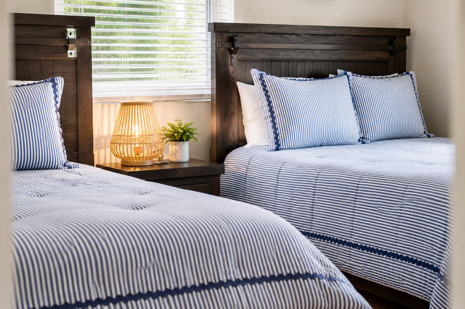A bedroom with two beds, each with a striped bedspread and matching pillows. There is a bedside table with a lamp and a small plant.