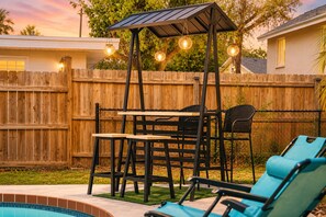 Une terrasse extérieure avec une table et des chaises en bois, une piscine et un espace salon couvert agrémenté de guirlandes lumineuses.