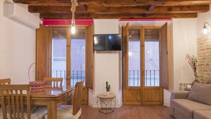 Een woonkamer met een houten eettafel, stoelen, een bank, een aan de muur bevestigde televisie en een klein bijzettafeltje.