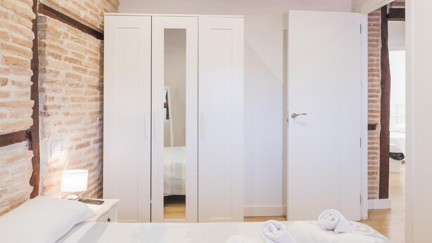 Quarto com parede de tijolos, cama com edredom branco, mesa de cabeceira com abajur e guarda-roupa com espelho.