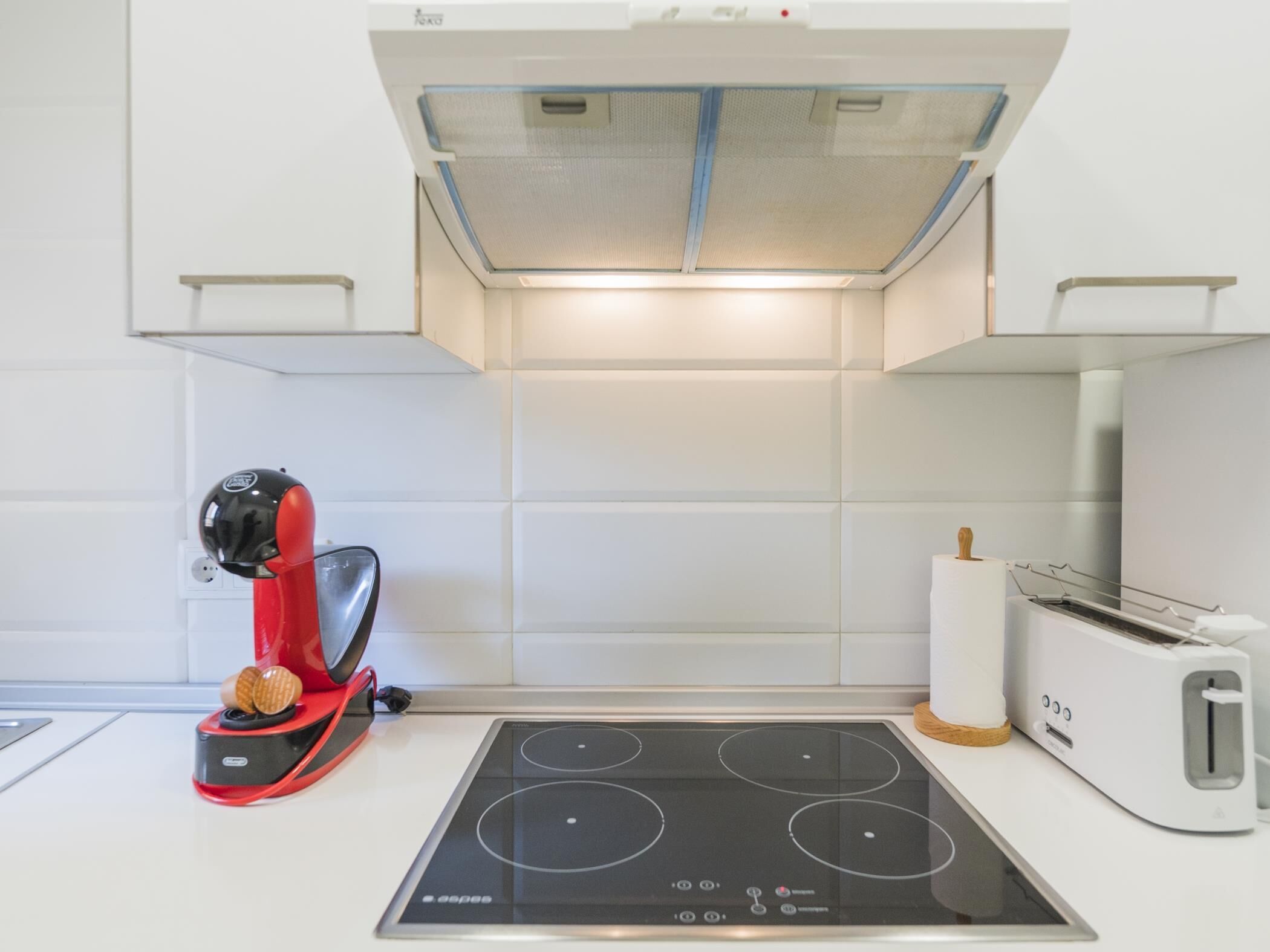 Una cocina con una hornilla de inducción moderna, un tostador de waffles rojo, un tostador y un salpicadero de azulejos blancos.