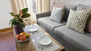 A dining table with plates, cutlery, and fruits, a potted plant, and a grey sofa with patterned cushions in a living room. - Collection Gran Vía (Madrid)