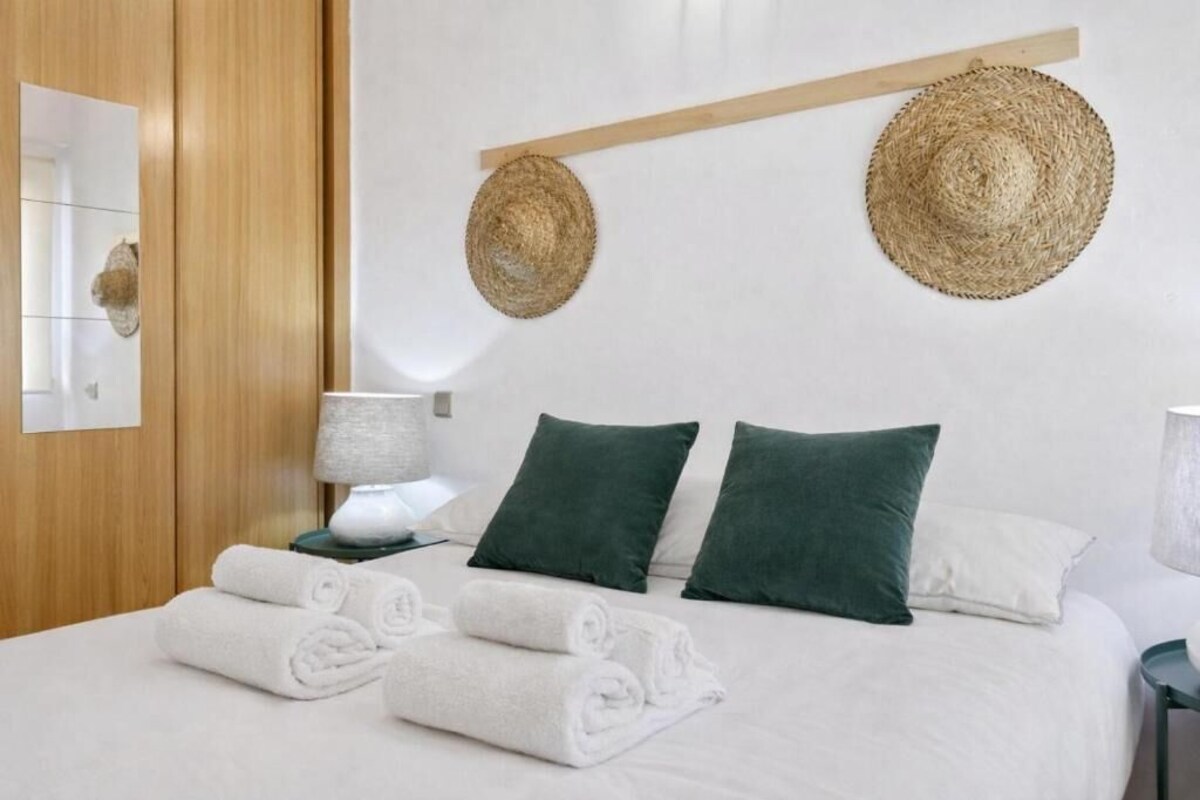 Habitación de hotel con una cama, dos almohadas, una mesita de noche con lámpara y dos sombreros tejidos en la pared.