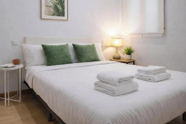 Una cama bien hecha con ropa de cama blanca y almohadas verdes, una lámpara de noche y una mesita con una planta.