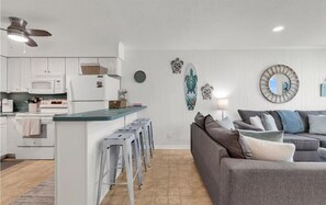 A modern kitchen with white cabinets, a central island, and bar stools. There is a gray sofa with multiple cushions and a round mirror on the wall. - Oceanfront Escape Shore Dr Condo Balcony (Myrtle Beach)