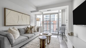 Ein modernes Wohnzimmer mit grauem Sofa, Couchtisch, Teppich, einem Gemälde und Blick auf Gebäude durch ein großes Fenster.