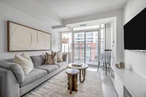 Un salon moderne avec un canapé gris, une table basse, un tapis, un tableau et une vue sur les immeubles à travers une grande fenêtre.