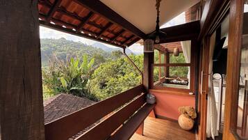 Varanda de madeira com um banco, luminária pendente e vista para vegetação exuberante e montanhas.