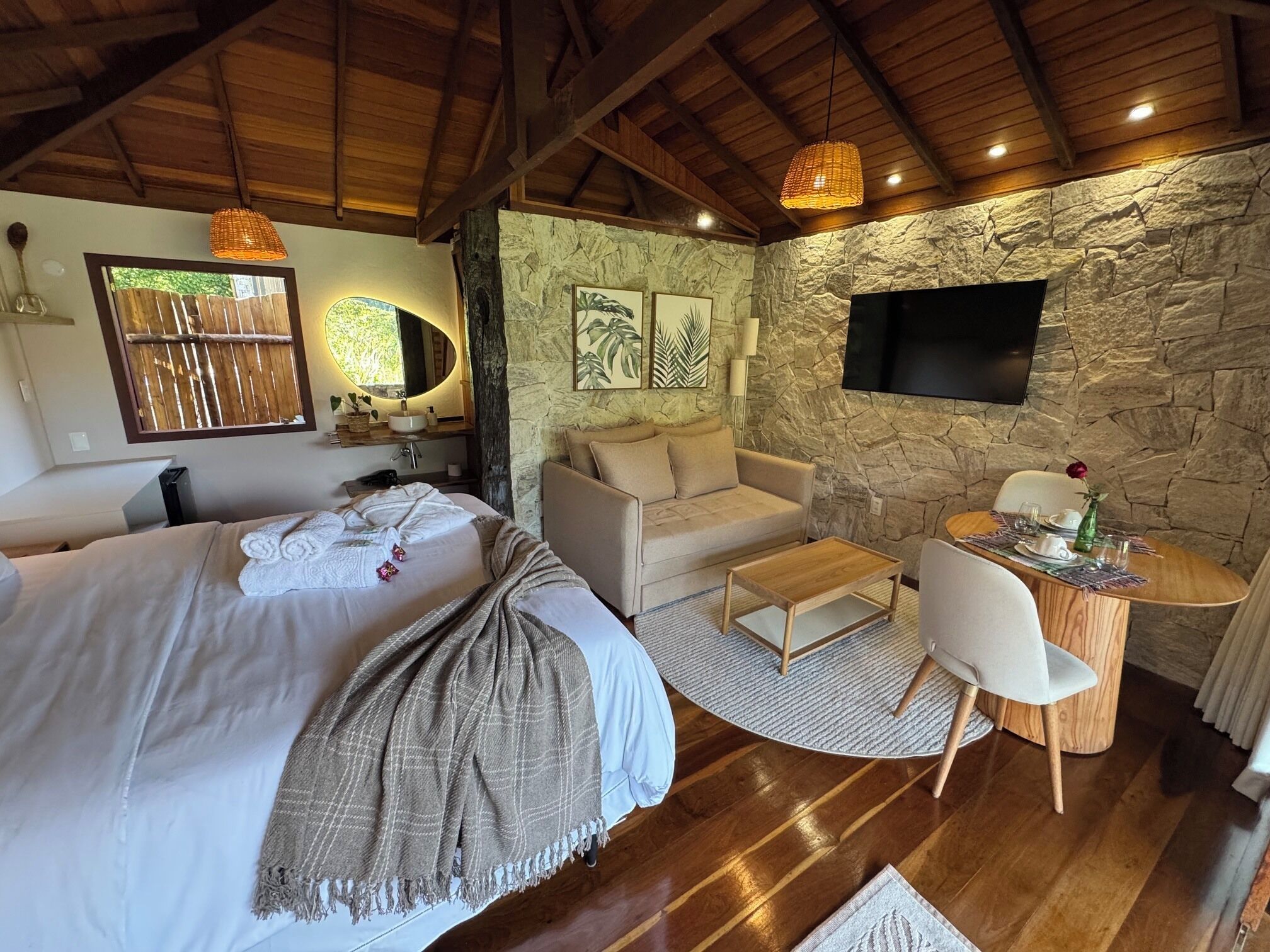 A bedroom with a bed, a sofa, a small table, and a TV mounted on a stone wall.