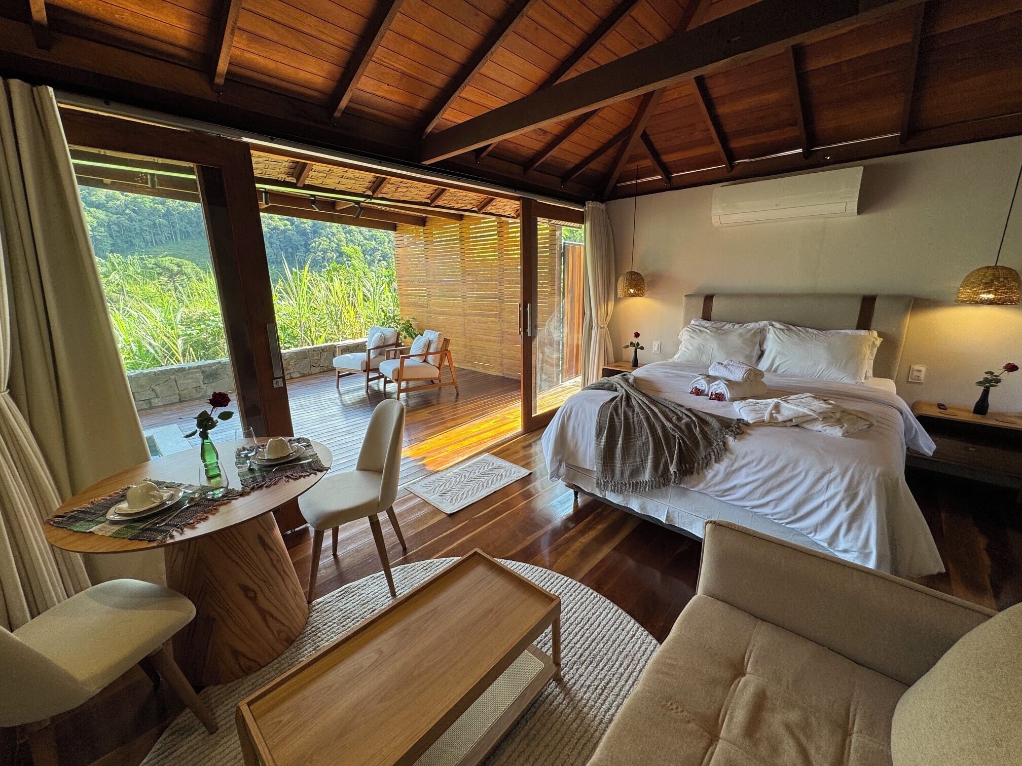 Un dormitorio con una cama grande, una zona de estar con sillas, una mesa de comedor y vistas a zonas verdes por las ventanas.