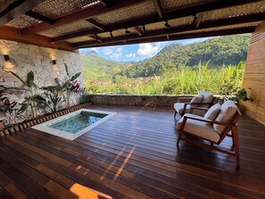 Una terraza de madera con piscina, dos sillas acolchadas y un muro de piedra con plantas.