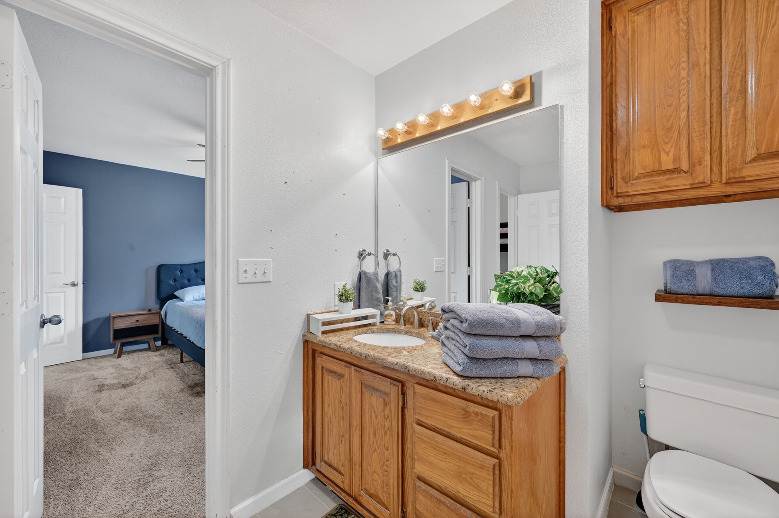 A bathroom with a wooden vanity, a sink, a mirror with lights, a toilet, and a blue wall.