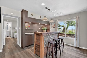 A modern kitchen with a bar area, wooden flooring, pendant lighting, and a view of the outdoors.