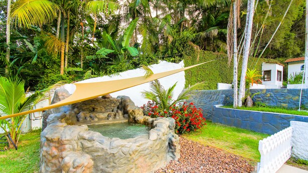 A stone hot tub surrounded by pebbles, with a white fence and tropical plants in the background. - Country house with heated pool, 7 bedrooms with air conditioning near São Paulo. (Juquitiba)