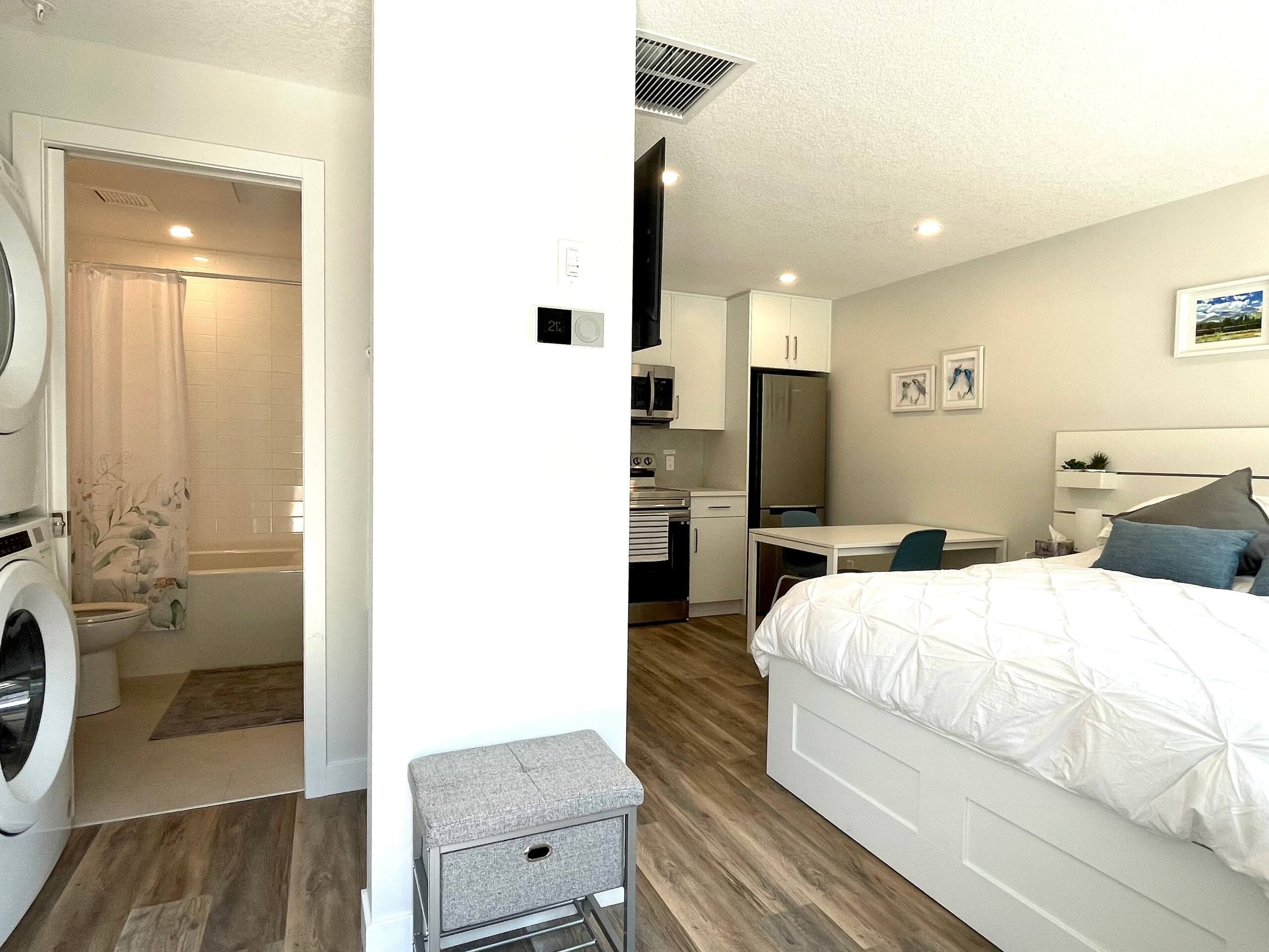 A compact hotel room with a bed, a desk, a washing machine, and a shower area.