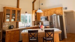 A kitchen with wooden cabinets, a central island with two chairs, a hanging light fixture, and a view of the outdoors. - Capay Valley Country Home (Guinda)