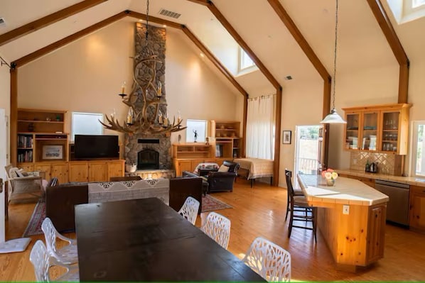 A spacious living area with a stone fireplace, wooden beams, a dining table, and a kitchen with a large island.