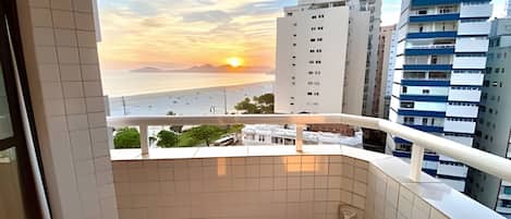 A balcony with a view of a sunset over the sea, adjacent buildings, and a tiled floor.