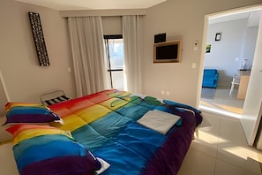 Un dormitorio con colcha colorida, una ventana con cortinas, un televisor montado y una puerta que da a otra habitación.