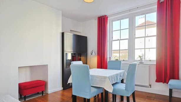 A dining area with a table and chairs, a red bench, a kitchen area, and a window with curtains. - Cosy Flat in Hammersmith - Close to tube station / But very quiet location (London)