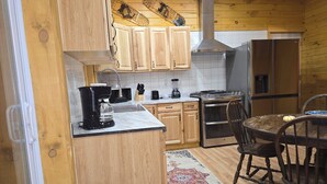 A kitchen with wooden cabinets, a stove, a refrigerator, a coffee maker, and a dining table with chairs. - Peaceful 2-bedroom cabin with AC in charming Irons, hot tub, sauna, covered deck (Irons)