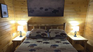 A wooden cabin room with a bed featuring a buffalo print quilt, two bedside lamps, and a framed picture on the wall. - Peaceful 2-bedroom cabin with AC in charming Irons, hot tub, sauna, covered deck (Irons)