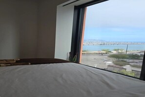 A room with a bed, a window with a view of a cityscape and water.